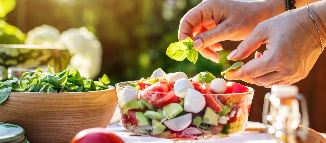 Female hand finishing fresh vegetable salad with herb basil leaves. Preparing healthy food in garden outdoors
