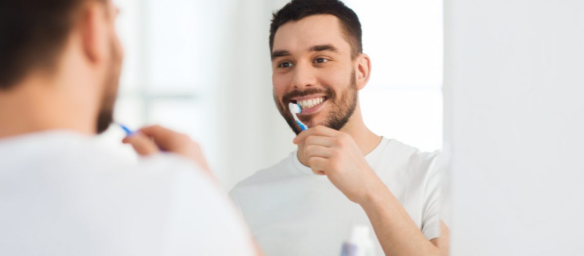 health care, dental hygiene, people and beauty concept - smiling young man with toothbrush cleaning teeth and looking to mirror at home bathroom